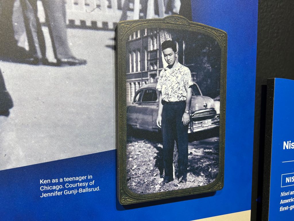 Close up of a photo on a blue museum panel. The photo is black and white and depicts a Japanese American male teenager standing in a patterned shirt and black pants, standing in front of a car. 