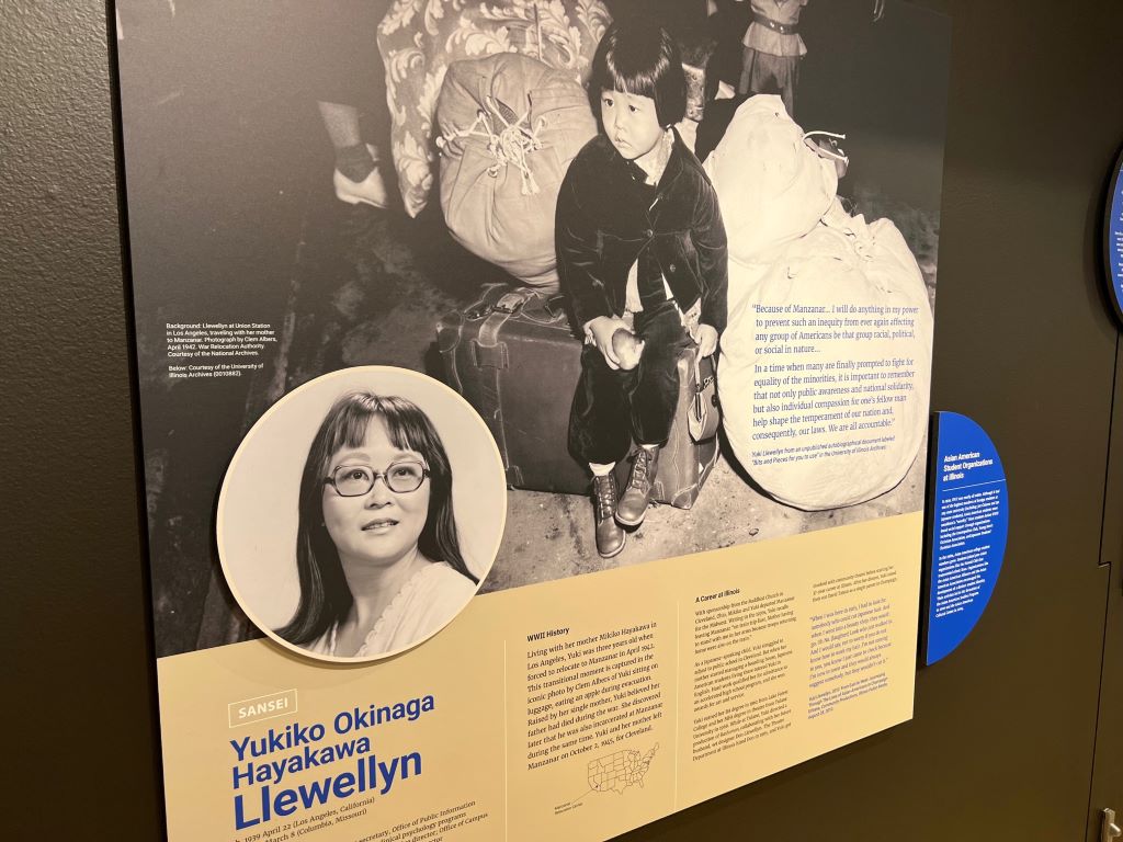 A museum panel that features a large image of a young Japanese American girl sitting on a piece of luggage. In a circle below is an image of a Japanese American woman in glasses. Below the images there is a block of text on a yellow background.