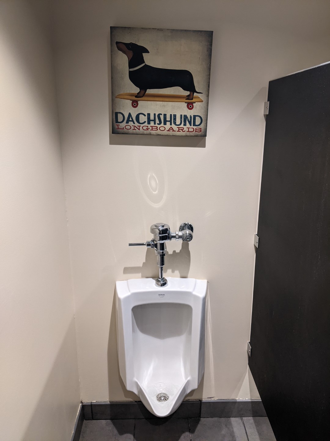 A urinal hangs on a beige wall. Above it is a painting of a dachshund on a skateboard with the words Dachshund Longboards.