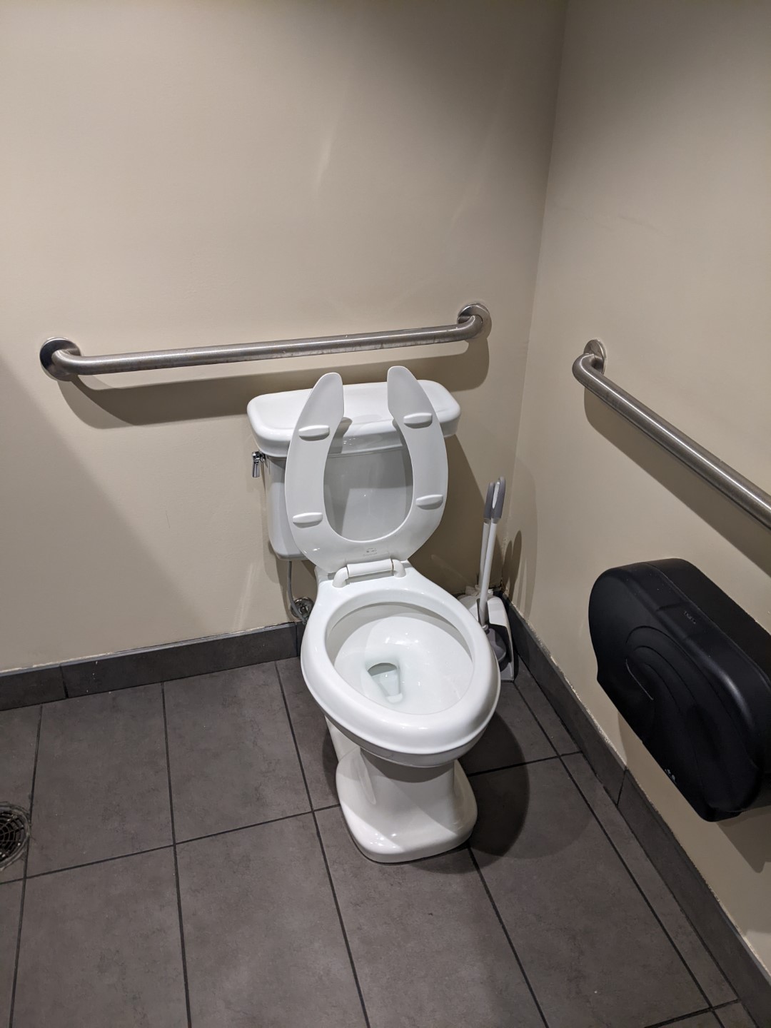 A white toilet with the seat lifted up. It's surrounded by beige walls, and it's on a dark gray ceramic tiled floor. There are guardrails on each wall. 