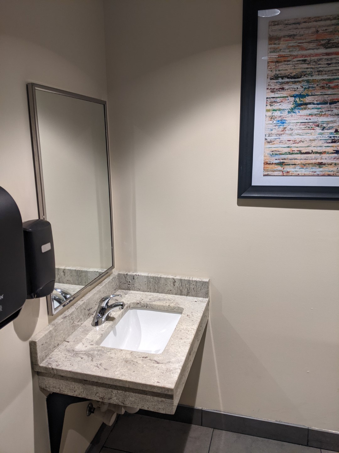 A marble sink with a white basin. It's attached to a beige wall and there's a mirror over it. A piece of artwork in a black frame hangs on the adjacent wall.