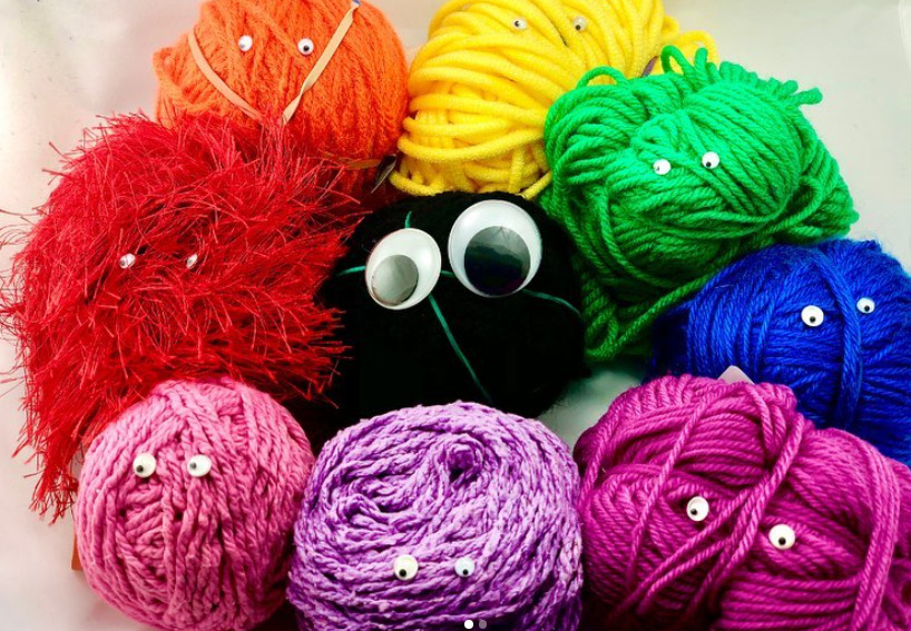 A black ball of yarn has big googly eye stuck to it. It is surrounded by colorful balls of yarn with tiny googly eyes.
