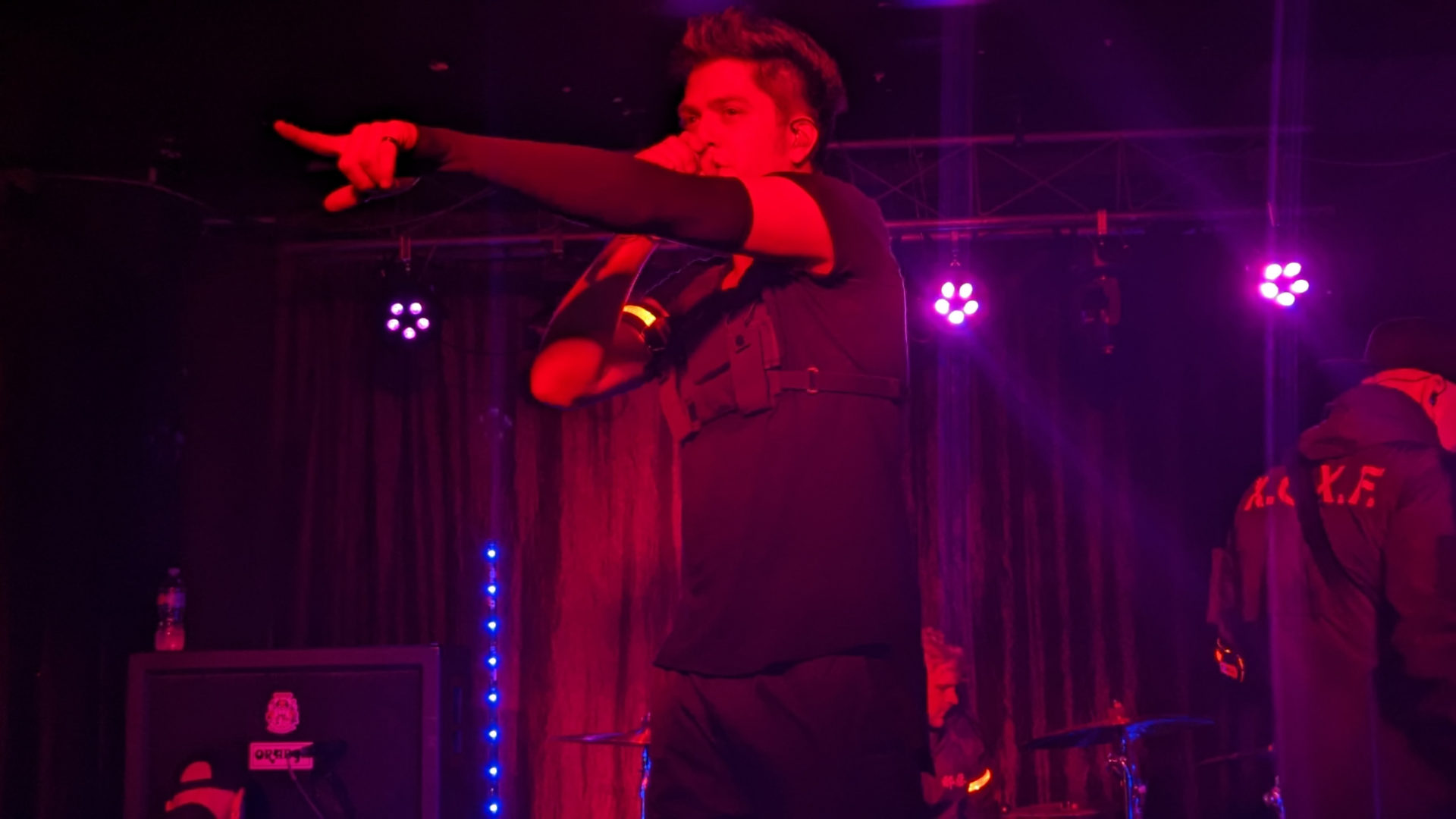 The lead singer of the band Versus Me, James Milbrandt on stage during a performance at the Canopy Club. He is wearing all black, with a black arm sleeve on his left arm. That arm is extended out, point to the crowd. He holds the microphone up to his mouth with his right hand. The lightings is red and pink and purple.