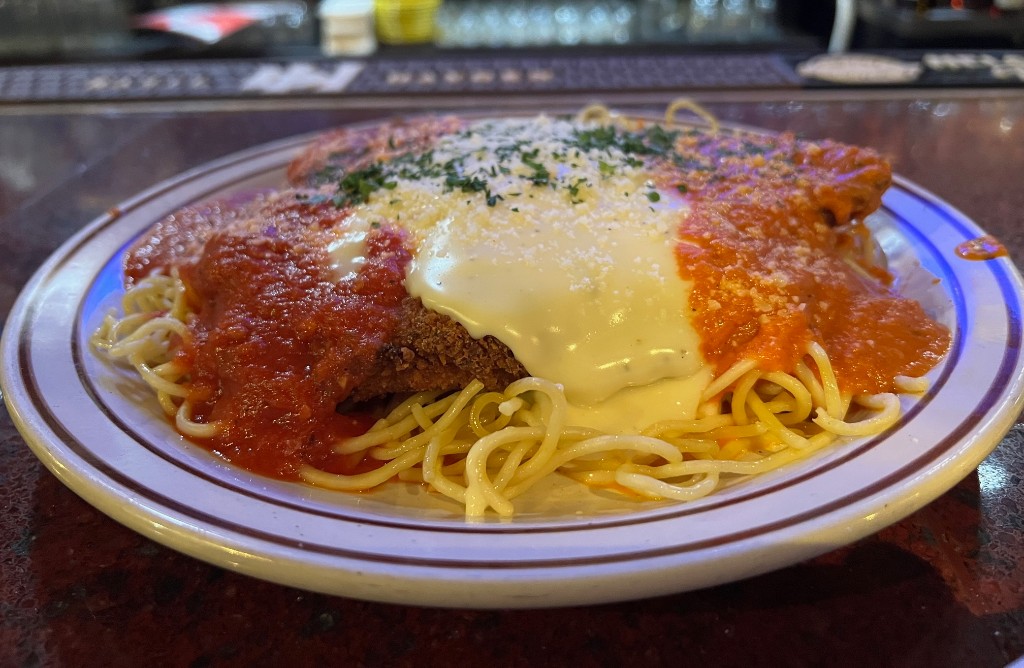 A round plated with spaghetti noodles, topped with a large piece of breaded chicken, and covered in a section of red marinara sauce, a section of melted mozzerella, and a section of orange vodka sauce. It's sitting on a wood bar.