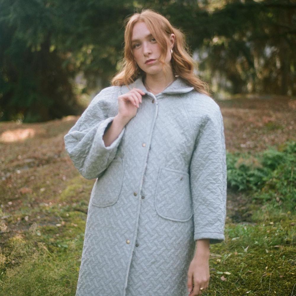 Bella White standing outdoors among trees. She is a thin, white woman with reddish hair. The photo is blurry to create an atmospheric effect. She wears a light blue quilted coat and uses her right hand to hold the collar of her jacket. 