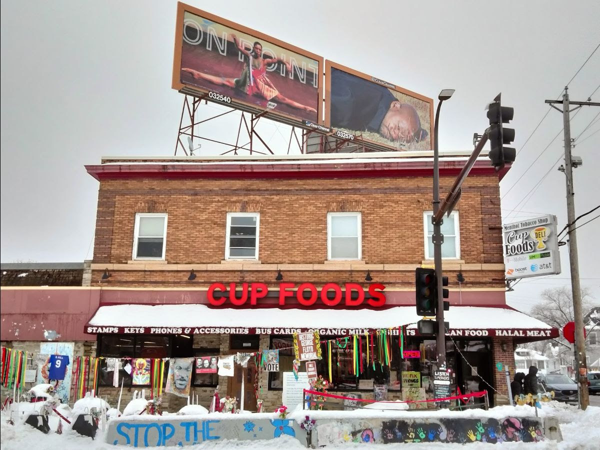 The exterior of a two story brick building. The words Cup Foods are in red on the front of the building. Above the roof are two billboards with artwork. In front of the building is a concrete barrier covered in graffiti messages and painted handprints. A clothesline hangs over it, with colored ribbons and other items hanging from it.