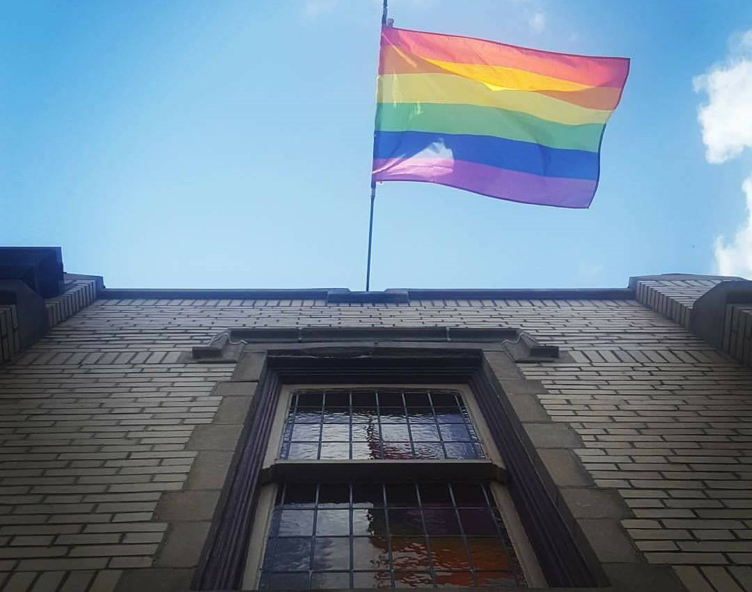 A rainbow flag juts out over a window on a stone building. There is blue sky behind the flag.
