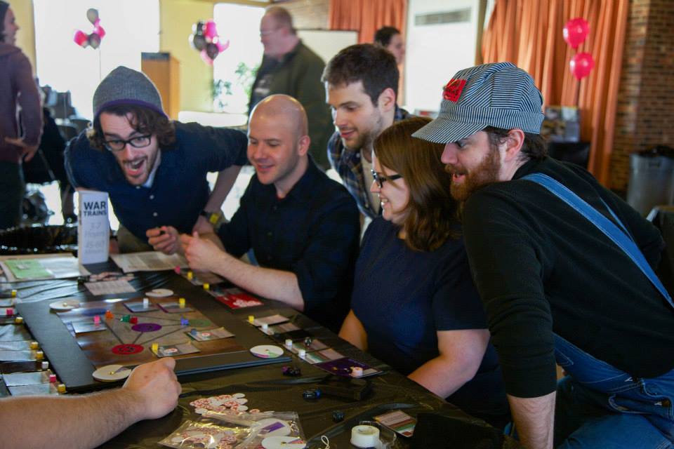A row of people are leaning over a board game on a table. they are all smiling or making surprised faces. 