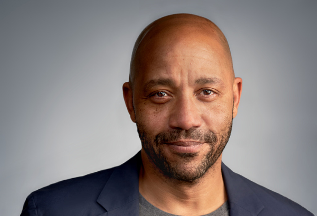 Headshot of a Black man who is bald with a closely trimmed mustache and beard. He was wearing a black suit jacket and gray shirt.