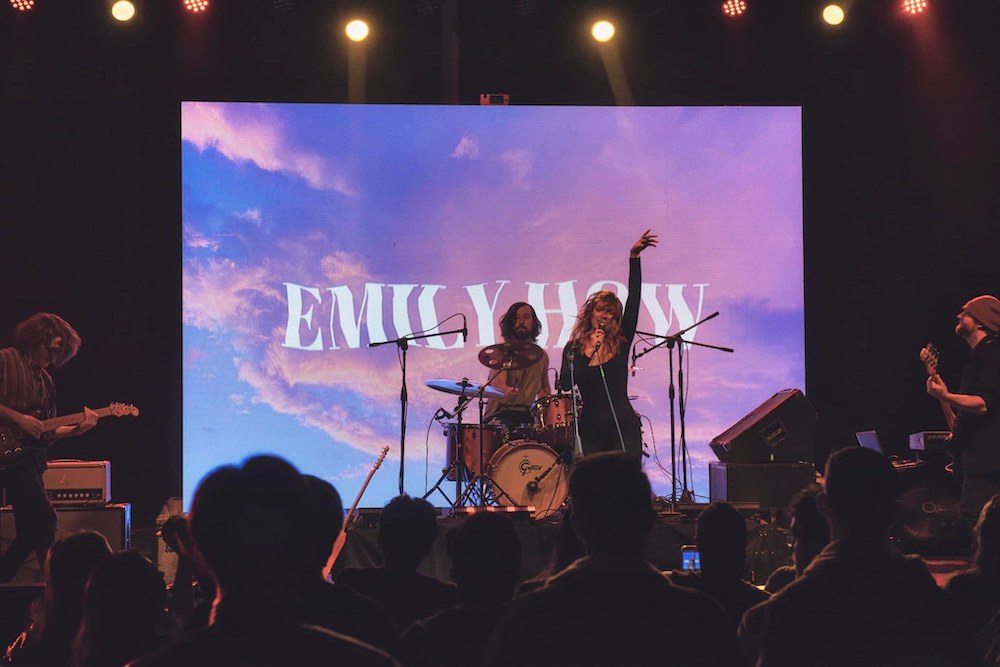 Musician Emily How on stage performing for a crowd. The bassist, guitarist, and drummer are all men. A thin, blonde woman wearing a black dress is in the middle of the stage singing into a microphone. Her left arm is raised. The crowd is in front of the stage.