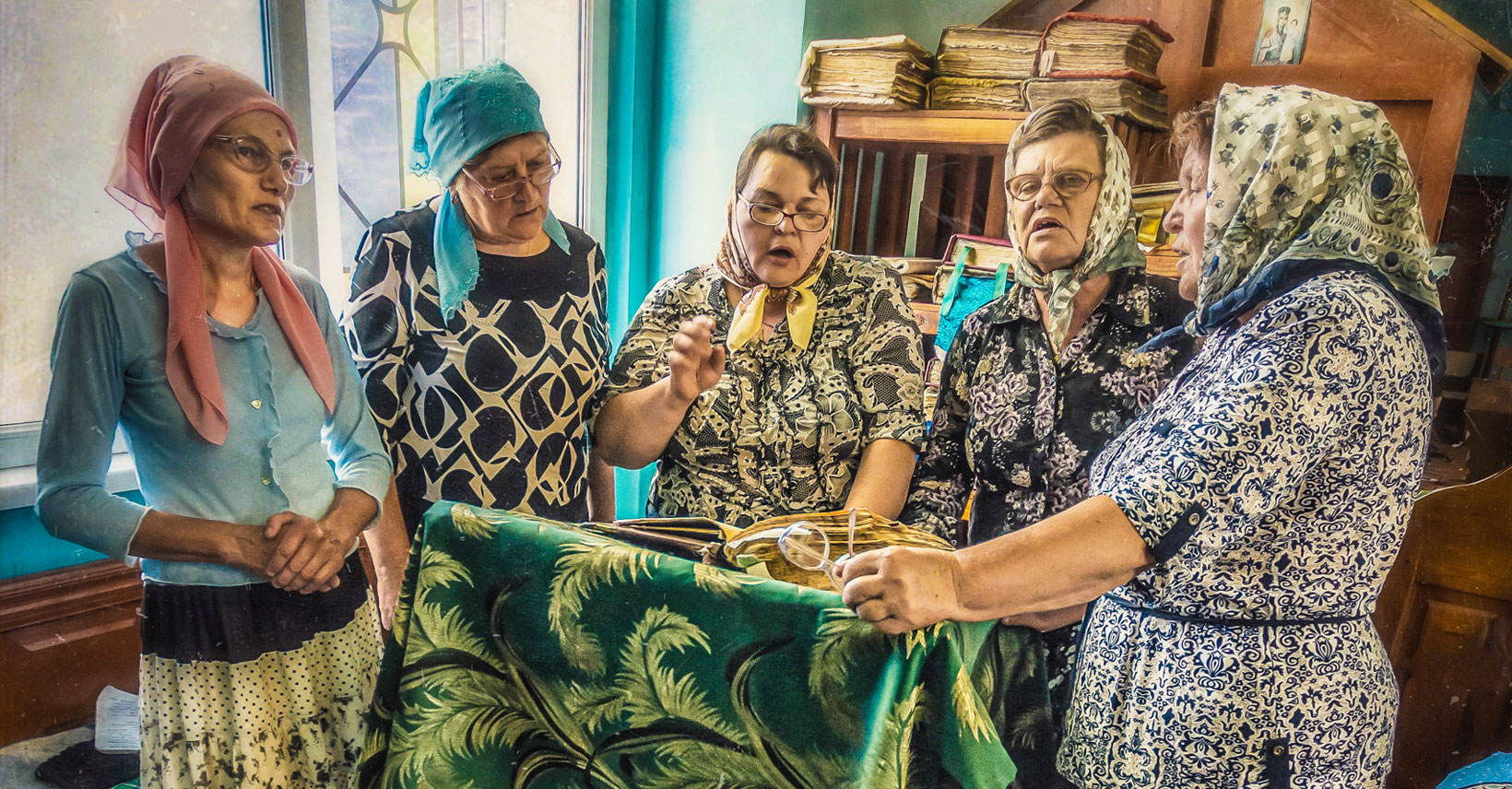 Five Ukrainian grandmothers are standing in semi circle around a lectern with an open book on it. It appears that they are reading or chanting along with the text. They all wear kerchiefs on their heads. Their clothes are colorful and patterned.