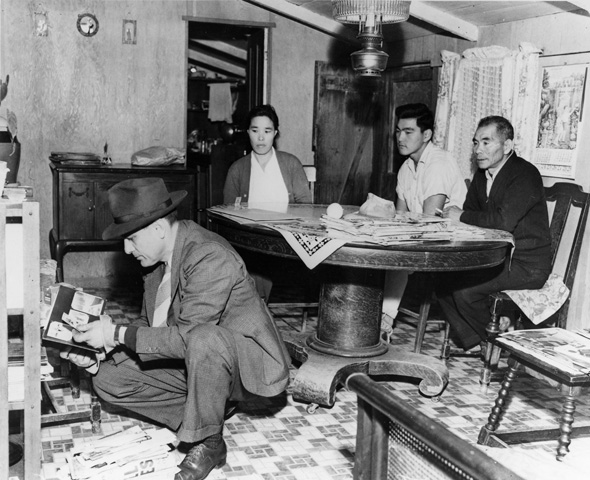 A black and white photo of three Japanese Americans sitting around a table, while a white man in a suit and hat is crouched down looked through books on a shelf.