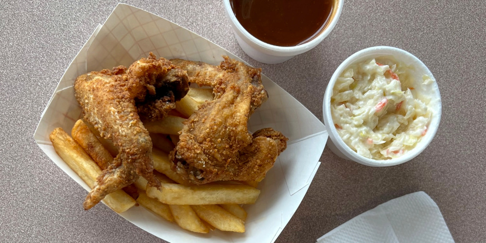 An overhead photo of fried chicken wings atop fries in a paper basket. Beside and cropped, there are two white cups, one with red barbecue sauce, and the other has coleslaw at Black-owned restaurants Wood N' Hog Barbecue in Urbana, Illinois. Photo by Alyssa Buckley.