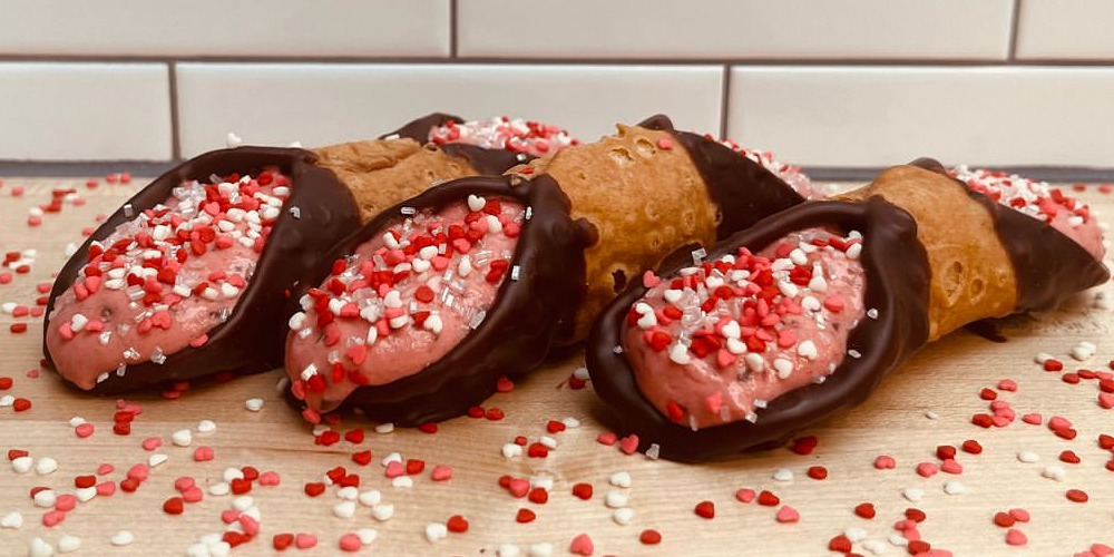 Three cannoli with chocolate dipped edges are filled with a pink strawberry cream with red and white sprinkles for Valentine's Day. They are on a wooden counter, and lots of sprinkles are spilled around them. Photo from Martinelli's Market on Instagram.