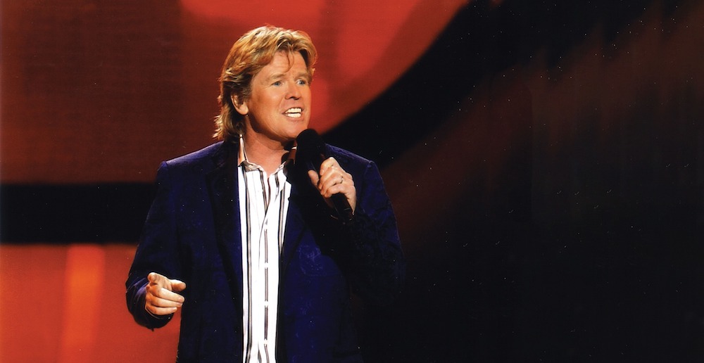 Peter Noone performs on stage, holding the microphone in front of a red and black background.