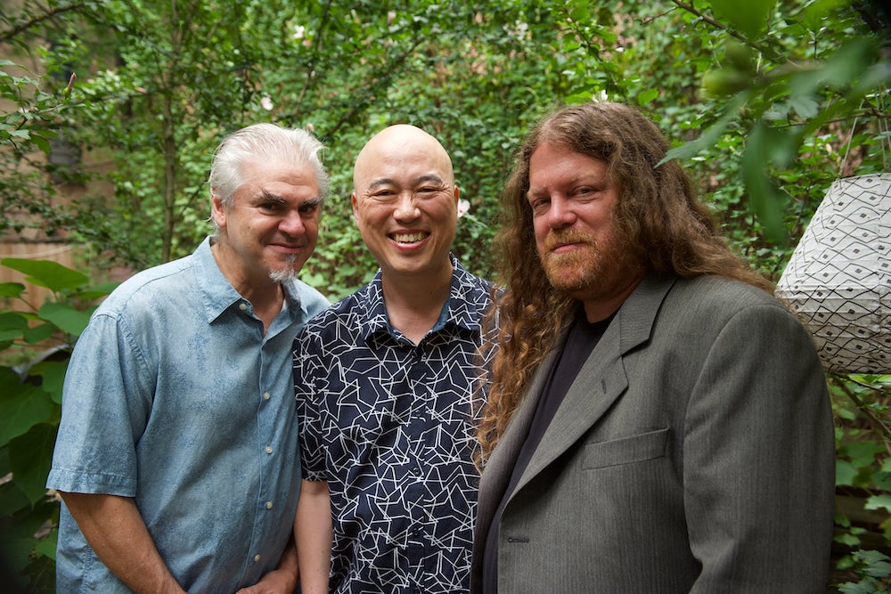 The Human Rites trio stands in front of a green, leafy backdrop outside, and smile for the camera. 