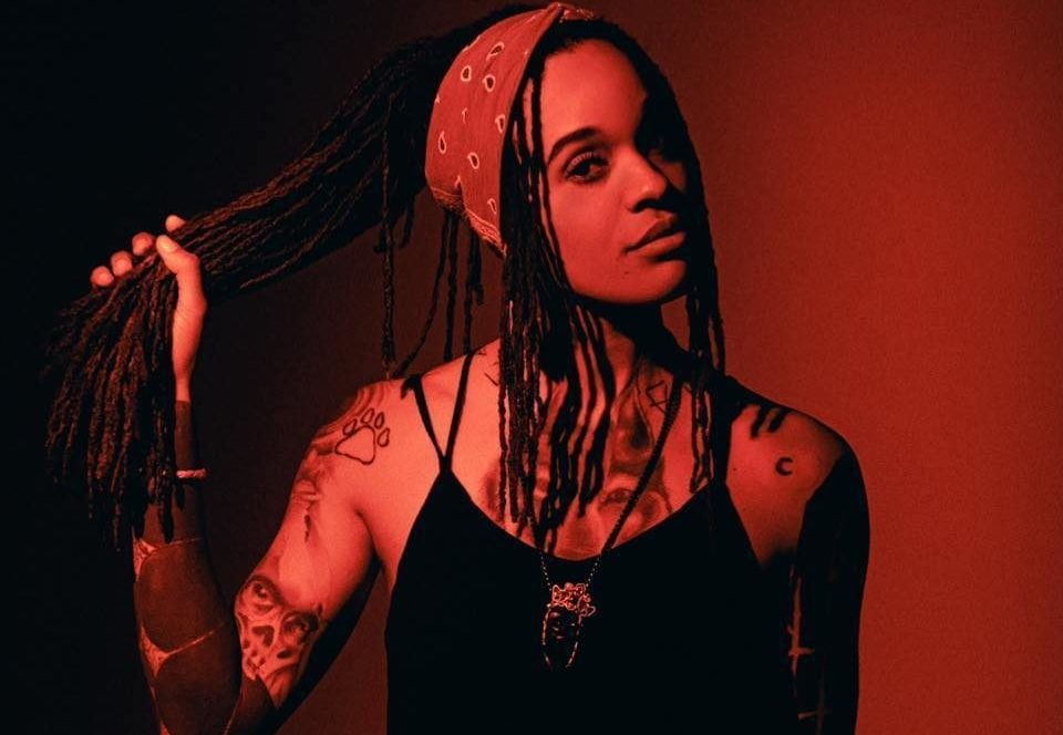 A light skin Black woman poses while holding a large portion of her long, braided hair in her right hand. She is wearing a black top with spaghetti straps and a reddish patterned bandana on her head. Her hair is long braids.The lighting is red and black.
