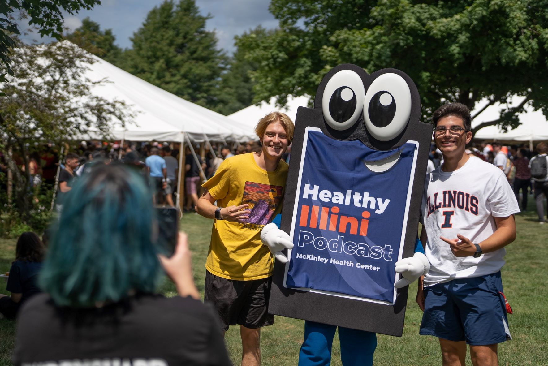 Image shows two smiling students posing with a cell phone character. Cell phone screen reads "Healthy Illini" in blue, orange, and white. A third student is in the foreground taking a picture.