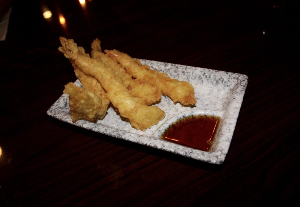 Four pieces of shrimp tempura at Sushi Siam. The breaded and fried shrimp are served on a rectangular plate, one corner of which contains a dipping sauce. The plate is a marbled black and white pattern. Photo by Rebecca Wells