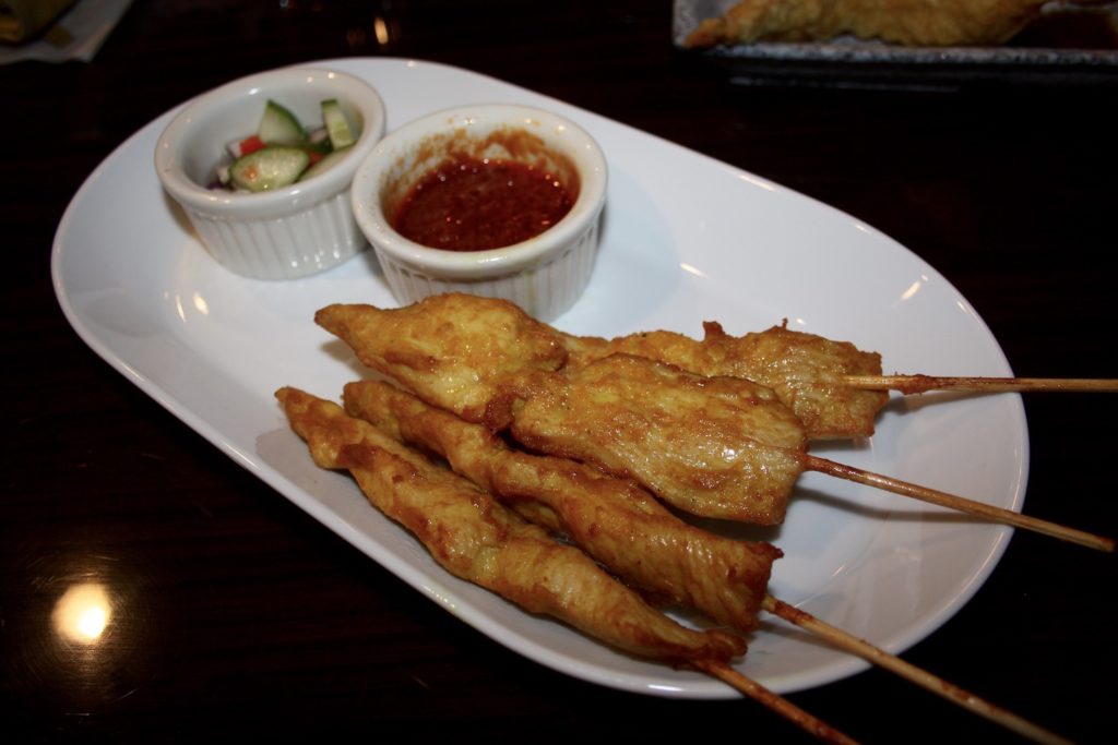 Chicken satay at Siam Sushi. Four skewers of golden brown cooked chicken are on an oval plate with two small condiment bowls, one with a peanut dipping sauce, the other with a cucumber salad. Photo by Rebecca Wells.