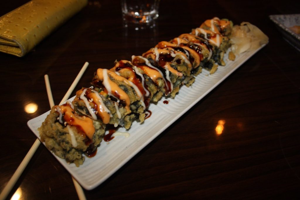 Tempura roll at Sushi Siam. The roll is served on a long white rectangular plate. The roll is fried, and garnished with white, dark brown, and orange sauces. Light brown wood chopsticks are on the left side of the plate. Photo by Rebecca Wells