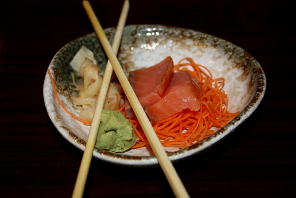 Smoked salmon sashimi at Sushi Siam. A small round bowl hold pickled ginger, wasabi, thinly sliced carrot, and two pieces of pink salmon. Light colored wood chopsticks rest across the bowl. Photo by Rebecca Wells.