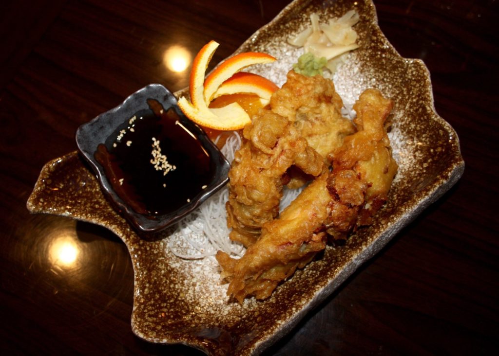 Soft shell crab at Sushi Siam. The fried crab is served on a brown and white asymmetrical plate. On the plate is a small, black bowl with dipping sauce, pickled ginger, and a small dollop of wasabi. An orange peel is fanned out as garnish. Photo by Rebecca Wells