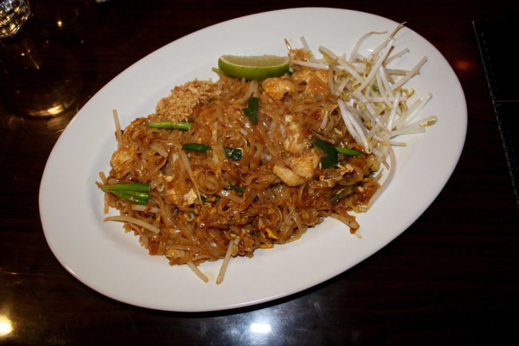 Pad Thai at Sushi Siam. An white oval plate holds brown stir fried noodles into which are pieces of scallions and chicken. A small garnish of crushed peanuts, a slice of lime, and some bean sprouts are also on the plate. Photo by Rebecca Wells.