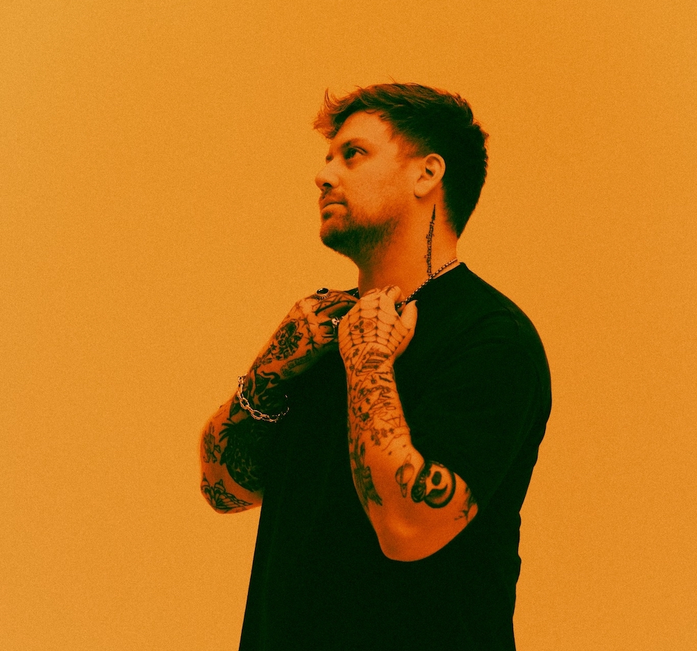 DJ Kayzo stands in front of an orange studio backdrop, he holds the chain around his neck, and looks off in the distance. 