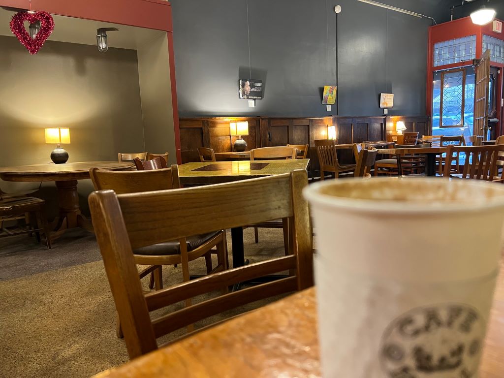 A close up of a white to-go cup of coffee. The background is a dark blue-gray wall with brown chairs and tables scattered throughout.