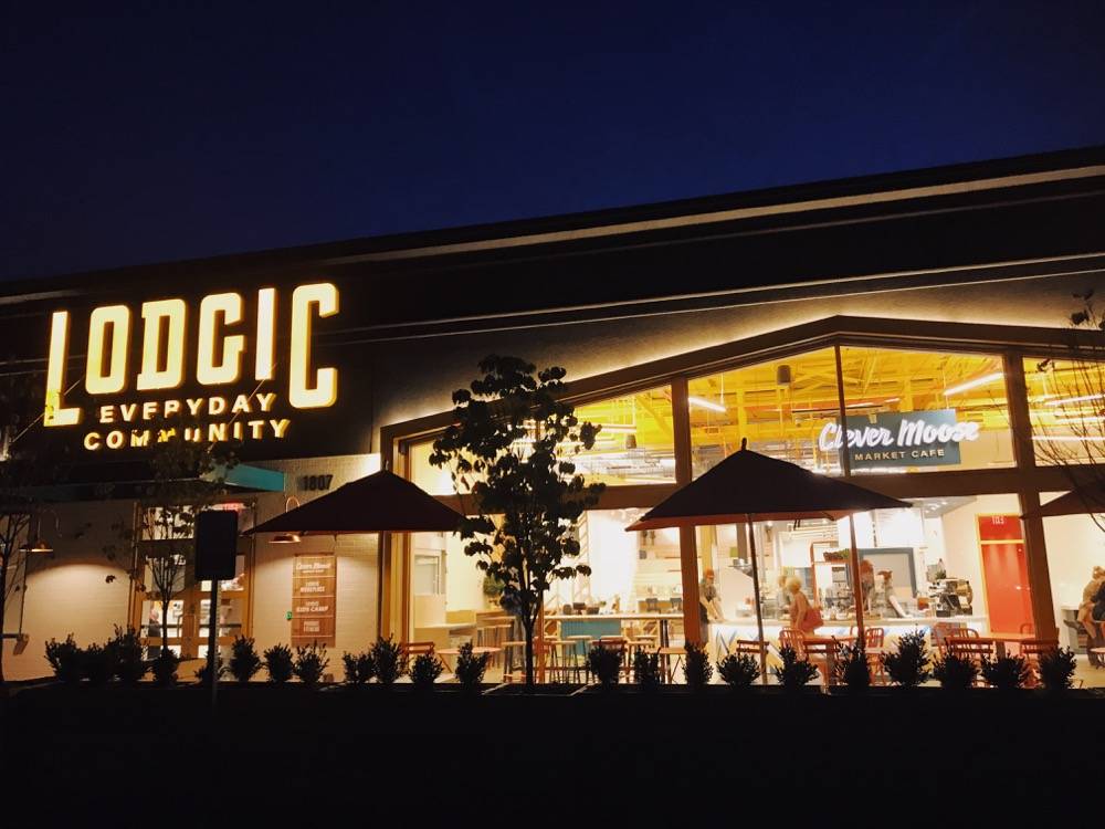 The front entrance of a business, at night. The facade is mostly windows, and there's a glowing yellow sign that says Lodgic in large block letters, and Everyday Community in smaller letters.