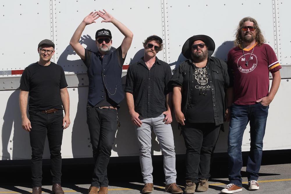 Miles Nielsen and his bandmates pose in front of what looks like the side of a semi truck. They look into the camera with serious looks on their faces.