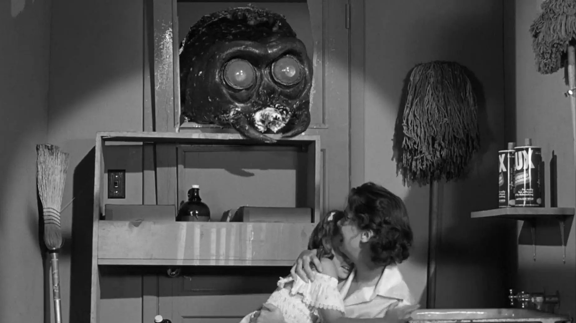 A black and white screenshot from a film. There is a large insect head poking through the window of a kitchen as a woman clutching her young child recoils.