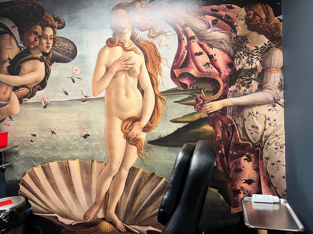 A wall is painted top to bottom with an image of a naked woman, covering portions of her body with her hand and her long red hair. She is standing in a shell, and there are people looking at her on either side. One is a woman in a light pink dress that is trying to cover her up with a large blanket.

A silver try and black chair sit in front of the wall.