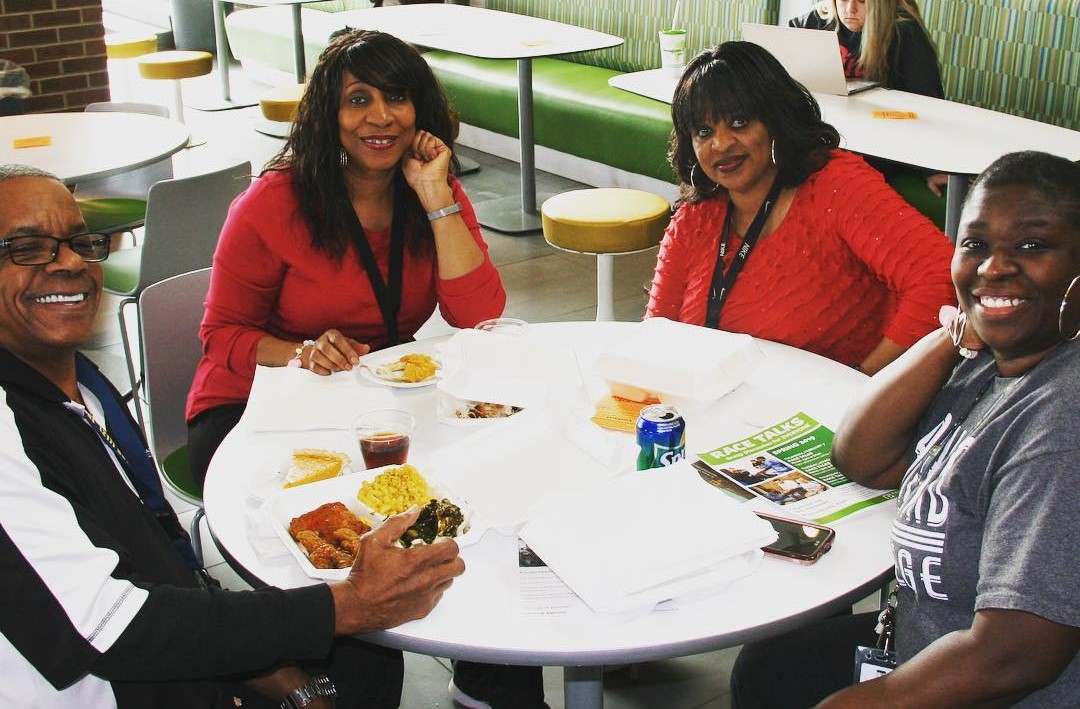 A Black man and three Black women are seated around a round white table. They have plates of food and papers in front of them.