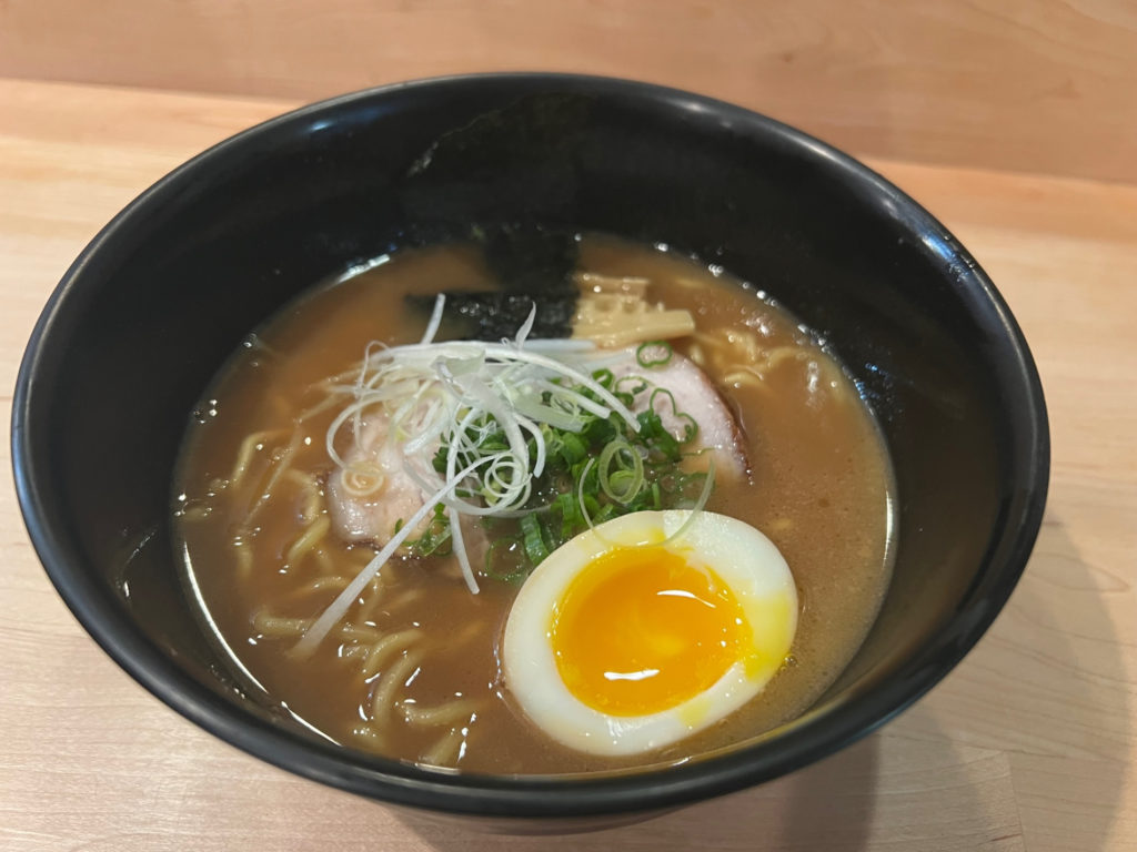A black bowl of ramen with half an egg in a light brown sauce.