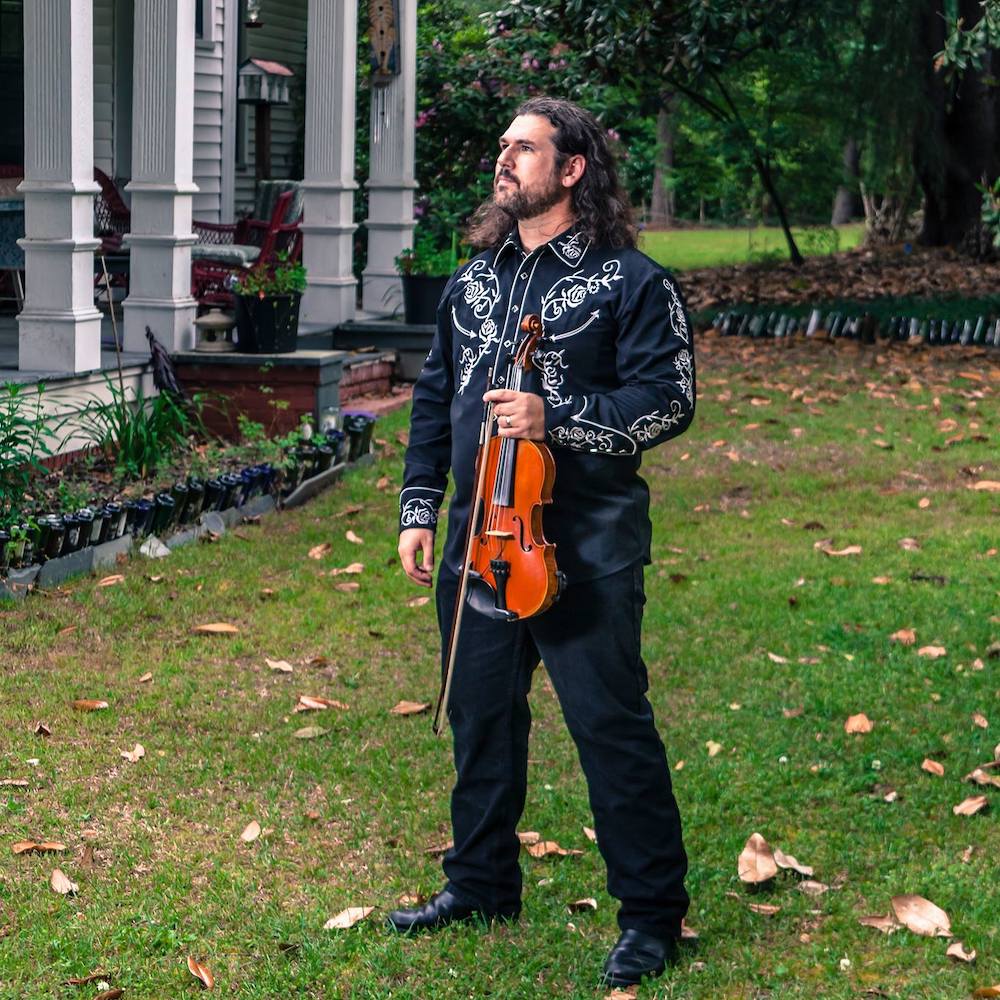 Dennis Stroughmatt stands outside in a yard, holding a fiddle and gazes off to the left of the screen. He has long curly hair and wears a black embroidered western shirt.