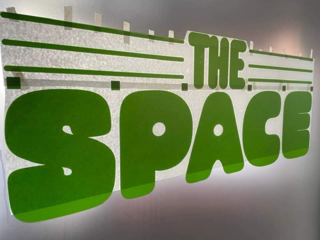 A sign with green bubble lettering is taped to a wall. It says The Space.