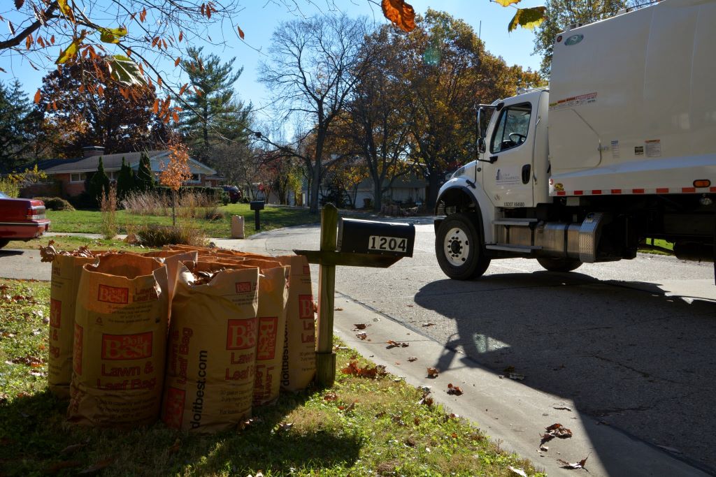 A white utility truck is parked by a curb. There are several paper lawn bags on the grass, next to a mailbox.