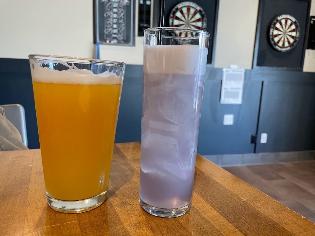 A picture of two drinks at Collective Pour in Champaign, Illinois. The one on the left is a beer in a pint glass. The one on the right is a light purple drink in a tall, thin glass. Dartboards are in the background. Photo by Remington Rock.