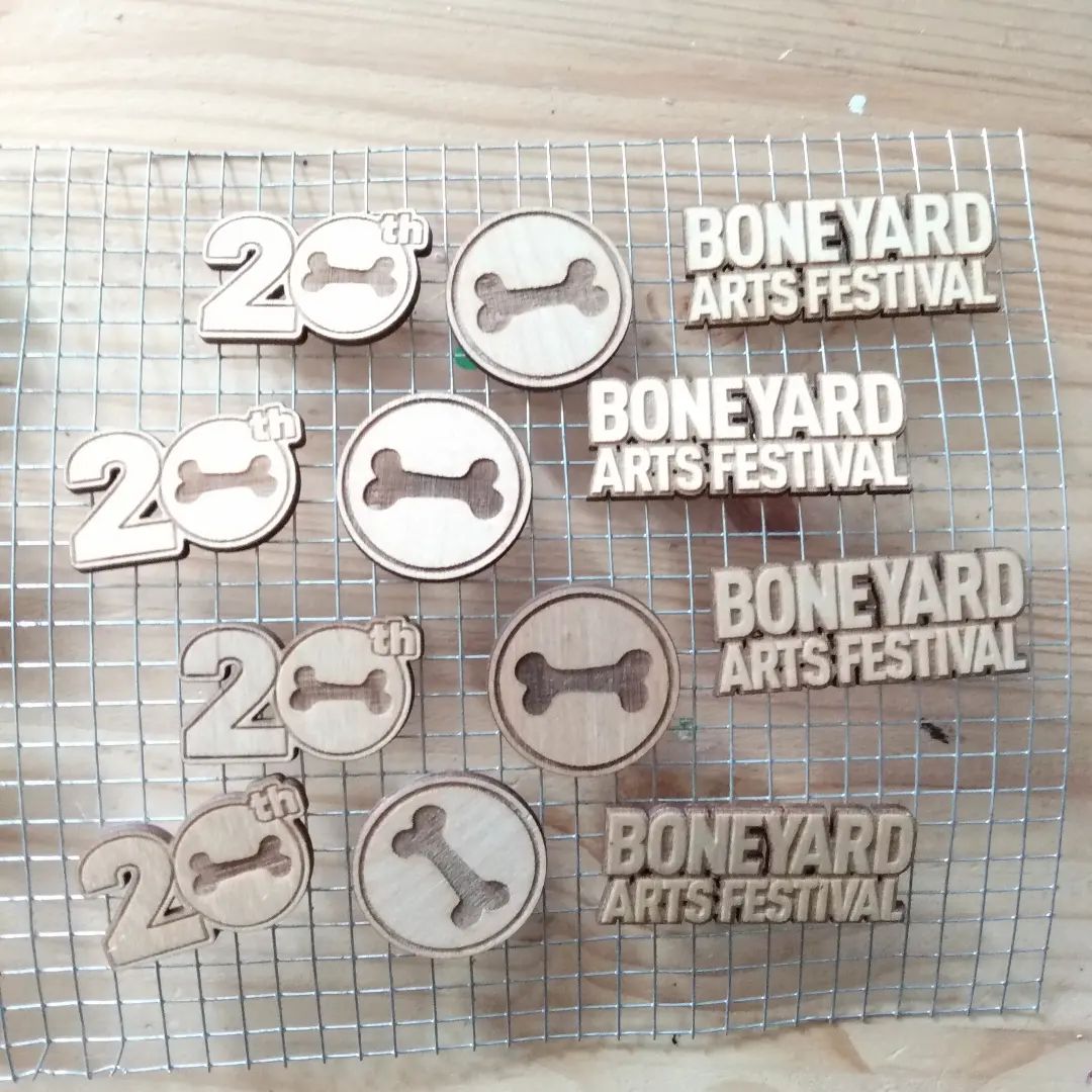 Wooden buttons that say 20th, Boneyard Arts Festival, and a circle with a bone in the middle are on a metal frame on a light wood table.