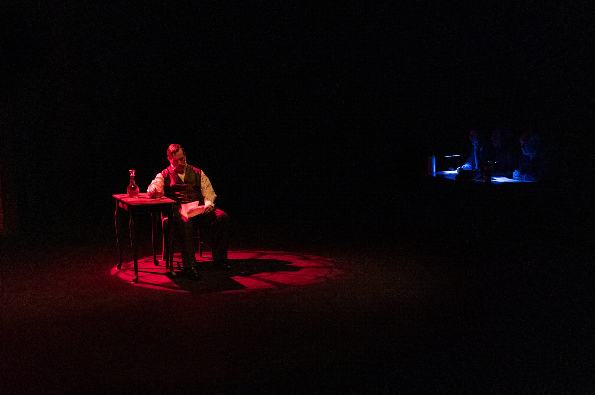 A dramatically lit theater stage. A red spot light is on a middle aged white man sitting at a desk reading a letter. In the background to the right of the image is a blue lit vignette.