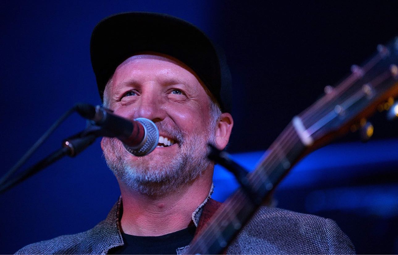 Photo of musician Miles Nielsen cropped from the shoulders up. He is wearing a black hat. He is has a gray beard and is smiling while playing guitar behind a microphone.