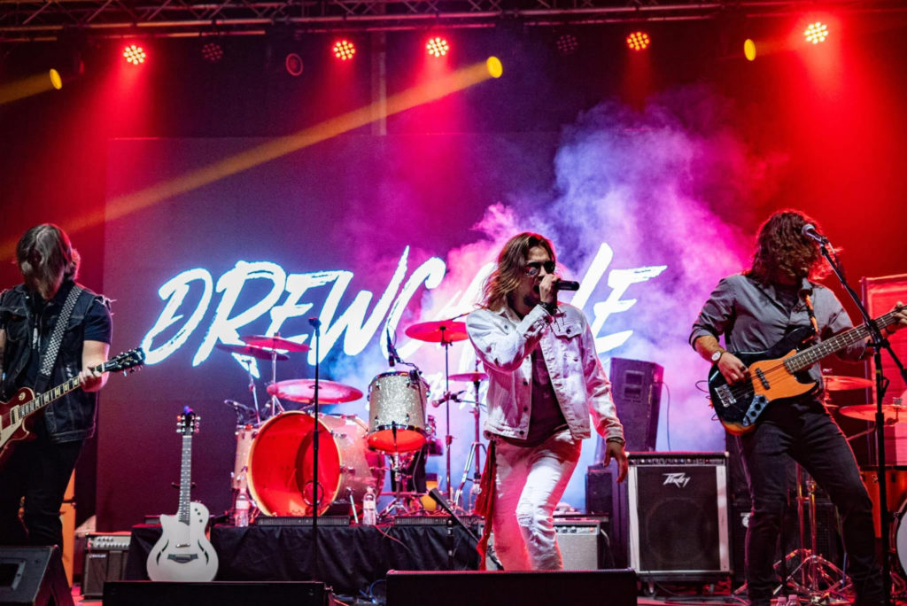 Drew Cagle & the Reputation on stage. The lights are red and there is a large "Drew Cagle" sign behind the band. Cagle, a white man, is in front wearing a ripped white jeans and a light colored jean jacket. He is wearing sunglasses and has long hair. There are two other white men pictured to his left and right playing guitar and bass. 