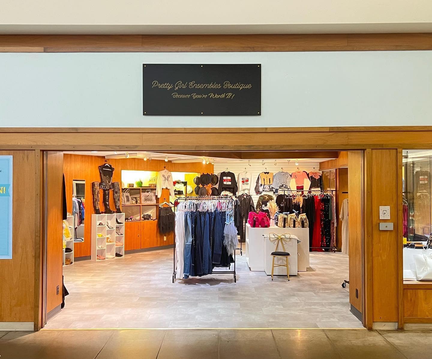 Image of a storefront surrounded by warm brown wood. The back white wall has a variety of jackets and in the foreground are racks of jeans and dresses. Shoes and accessories take up the left wall.
