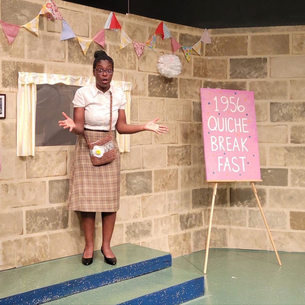 A Black woman stands with her hands extended in a questioning pose. She is standing in front of a faux brick wall, next to a sign that says "1956 Quiche Breakfast."