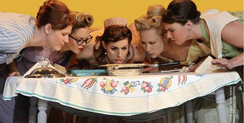Photo of five white women dressed in 1950s hair, makeup, and clothes leaning down staring into a series of quiches on table covered with a tablecloth covered in pictures of fruit.  