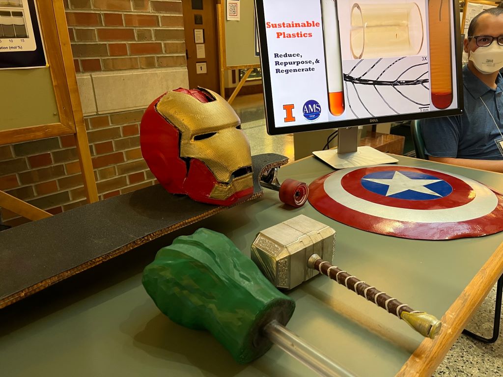 A shield, Ironman mask, club, and green  fist on a stick are sitting on a table. There is also a monitor that says Sustainable Plastics: Reduce, Repurpose, and Regenerate. 