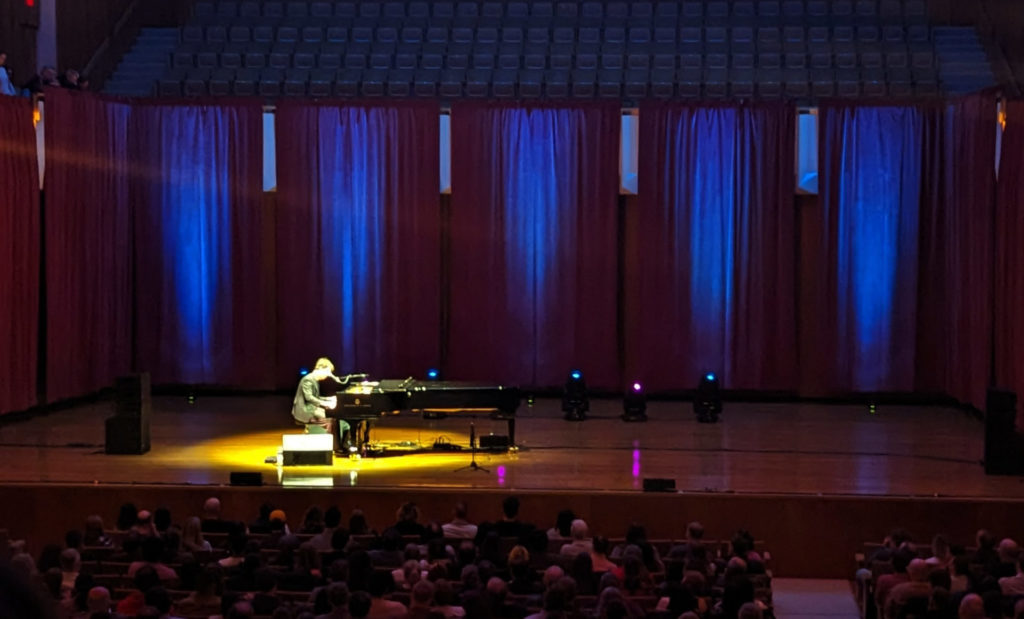Ben Folds alone on a stage under a spotlight playing a piano in a wide shot showing the width of the stage and the first 5 rows of people.