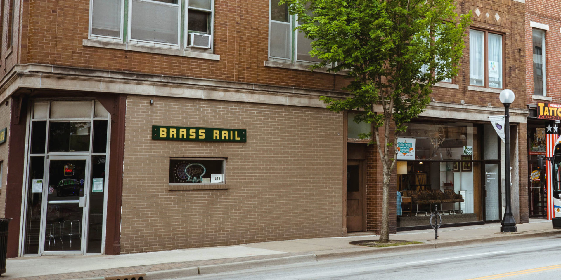 A cropped photo of Brass Rail in Downtown Champaign on a spring day. The exterior is brick, and there is a dark green sign with yellow letters reading the bar's name. Photo by Anna Longworth-Singer.